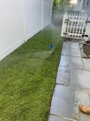 Narrow grass strip beside a white fence and tiled patio, with a small blue sprinkler spraying water.
