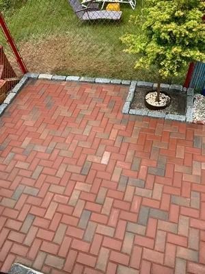 Red brick patio with a small tree in a stone border, bordered by grass and red wooden posts.