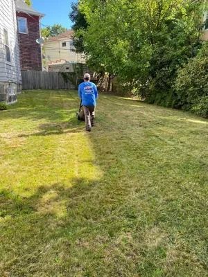 Person in a blue shirt mowing a grassy backyard with a push mower.