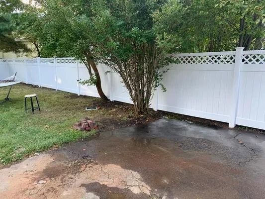 Wet backyard patio beside white fence and trees, with muddy puddles on the ground