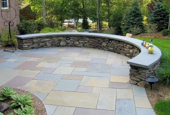 Curved stone garden bench with light paver seating area and potted flowers in a lush backyard.