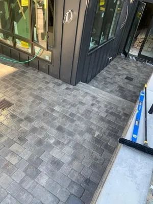 Paver walkway outside a glass storefront entrance with black trim and a blue rail on the right