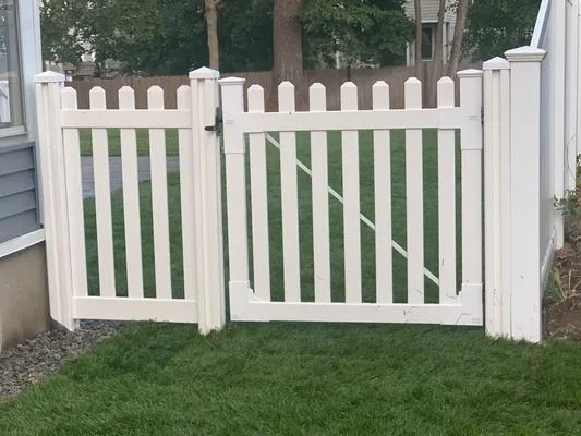 White picket double gate in a grassy yard with trees and a house in the background