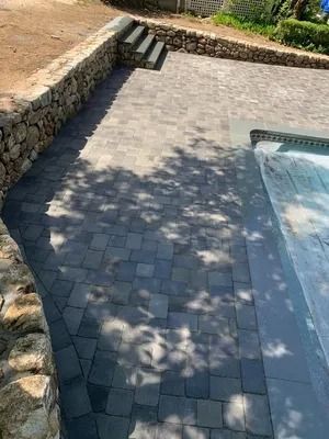Stone patio beside a pool, with steps and dappled tree shadows on gray pavers.