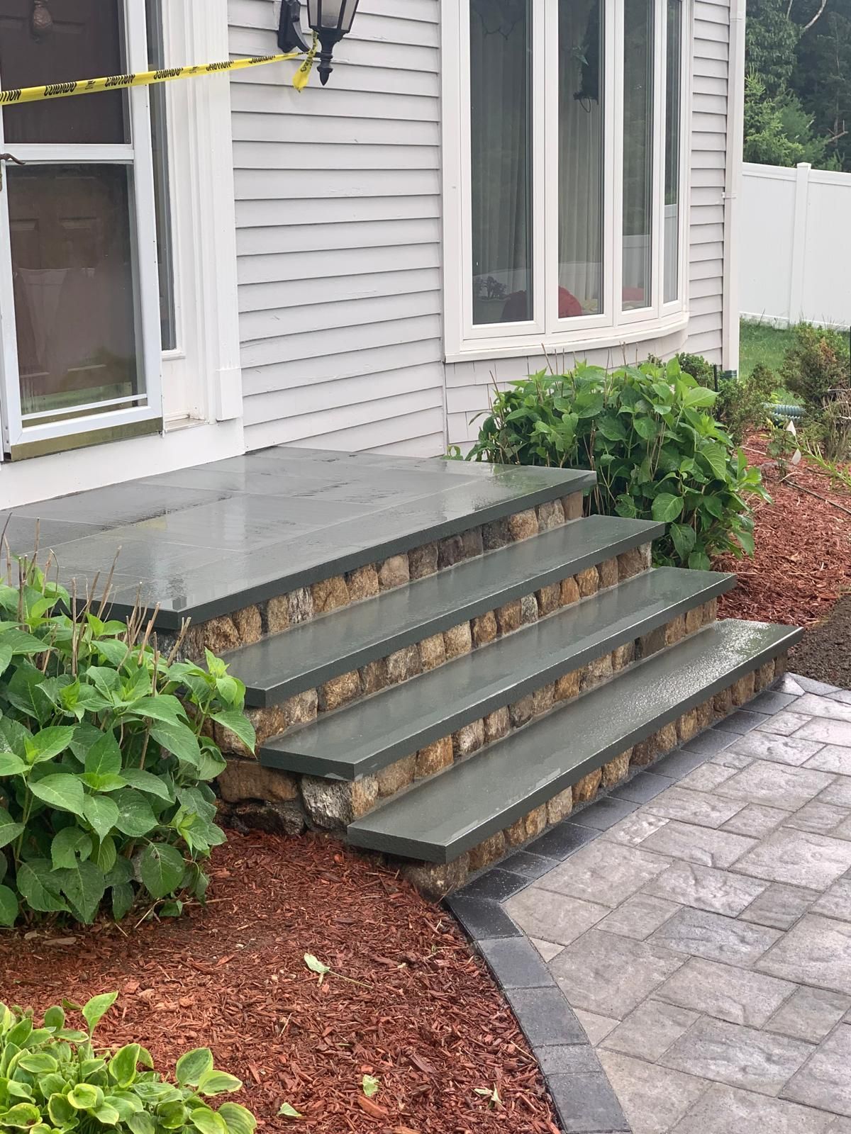 Front steps with gray pavers, a gray house facade, and green shrubs beside a walkway