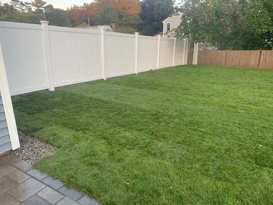 Backyard lawn bordered by a white privacy fence and a paved patio in the foreground