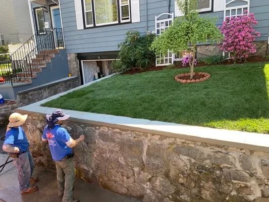 Two workers spraying a stone retaining wall below a blue house with a landscaped lawn and pink flowering tree.