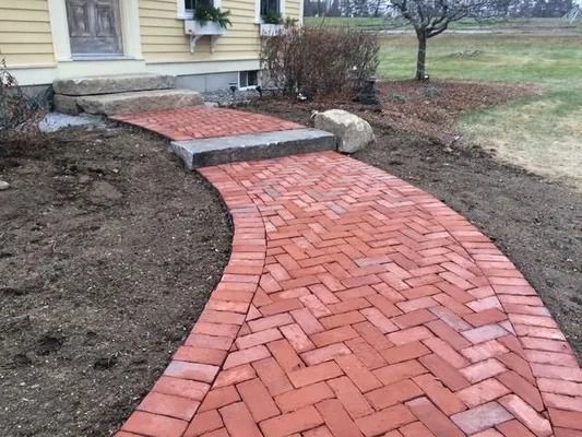 Red brick walkway curves to front steps of a house with newly landscaped soil and grass nearby