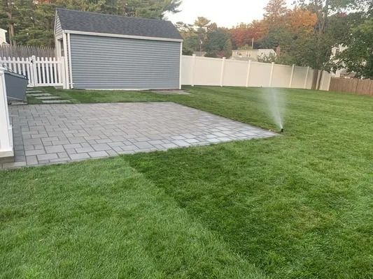Lawn sprinkler watering a green backyard beside a gray shed and paved patio