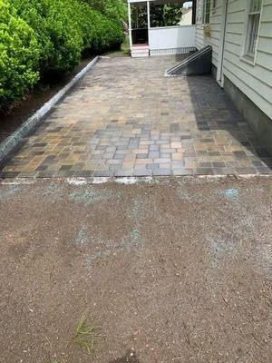 Stone paver patio beside a house, bordered by shrubs and a gravel driveway in the foreground