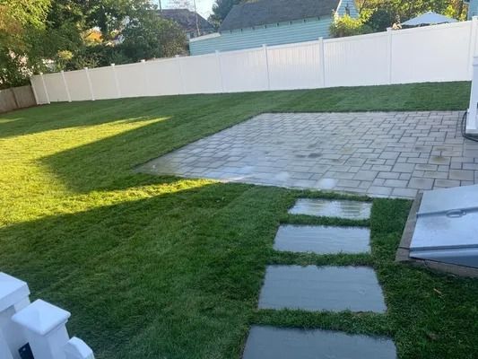 Backyard lawn with stepping stones leading to a paved patio beside a white fence