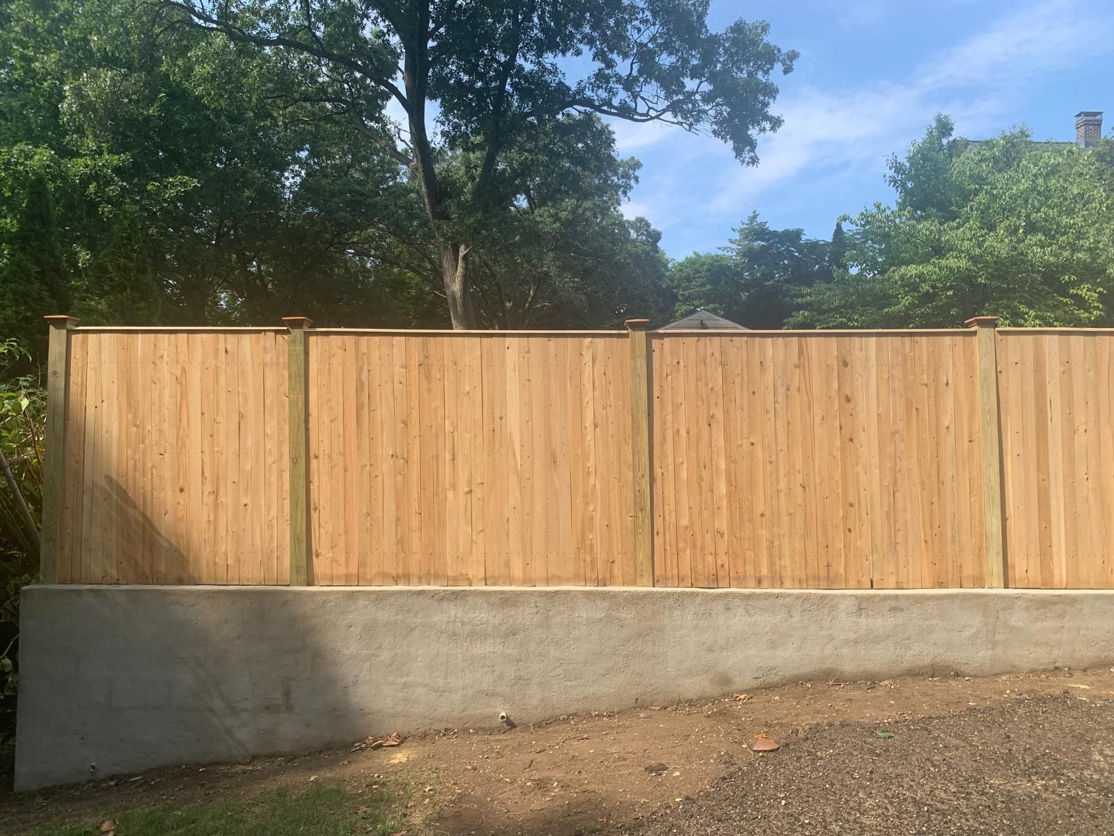 Wooden privacy fence above a concrete retaining wall, with trees and blue sky behind it.