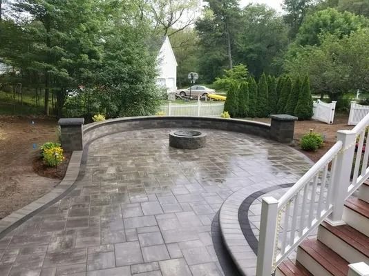 Curved stone patio with built-in bench and central fire pit, beside white stairs and lush trees.