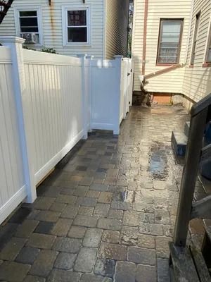 Narrow brick patio walkway beside a house, bordered by a white vinyl fence and small gate.