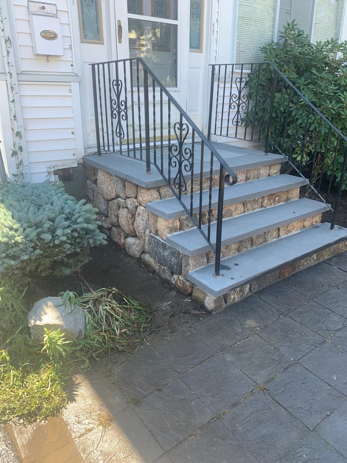 Front steps with black railing leading to a white house entrance, beside shrubs and a sunlit stone walkway.