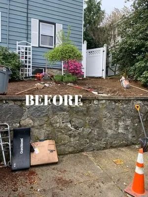 Before view of a house exterior with a stone wall, cone, yard debris, and a small dog near a white gate