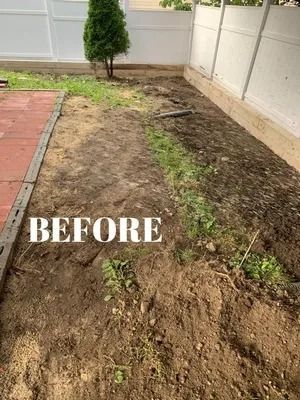 Bare dirt garden bed beside a brick path and white fence, with a small tree in the background.