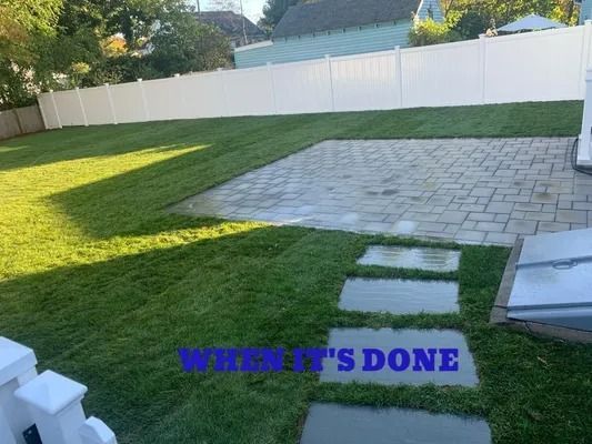 Backyard with green lawn, white fence, and a gray stone patio under sunlight.