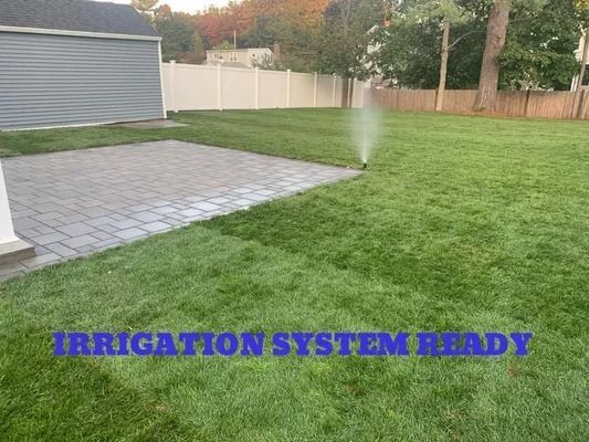 Backyard lawn with sprinklers watering near a paved patio and shed; text reads “IRRIGATION SYSTEM READY”