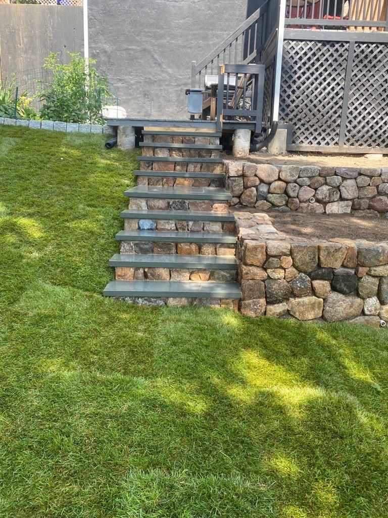Stone steps leading from a grassy lawn up to a wooden deck with lattice railing and stone retaining wall.