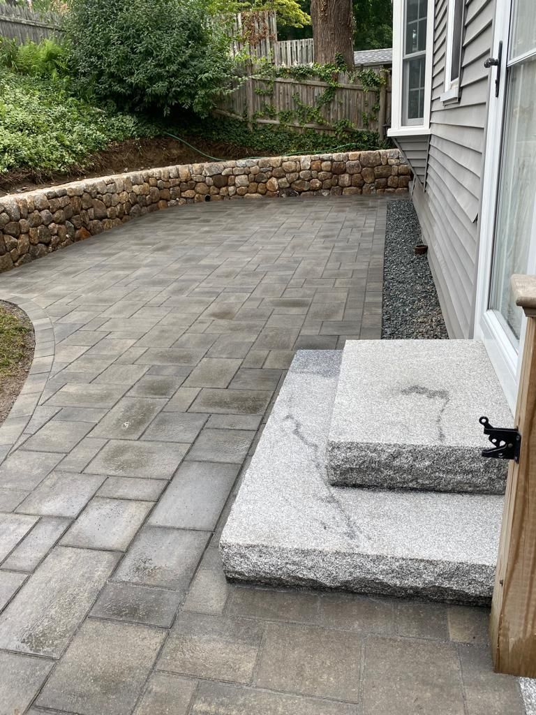Stone patio with curved retaining wall and two front steps beside a gray house.