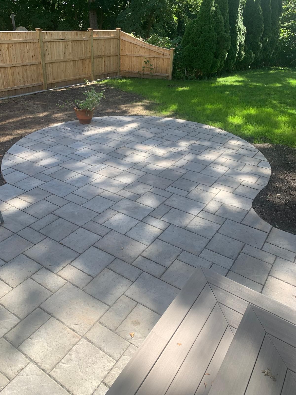 Backyard patio with a circular stone paver area, wooden fence, and lawn in sunlight