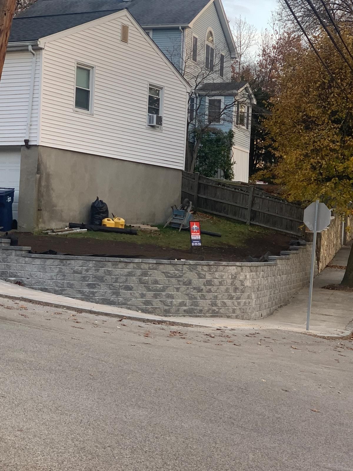 Residential yard with a stone retaining wall, white house, and a stop sign by the street.