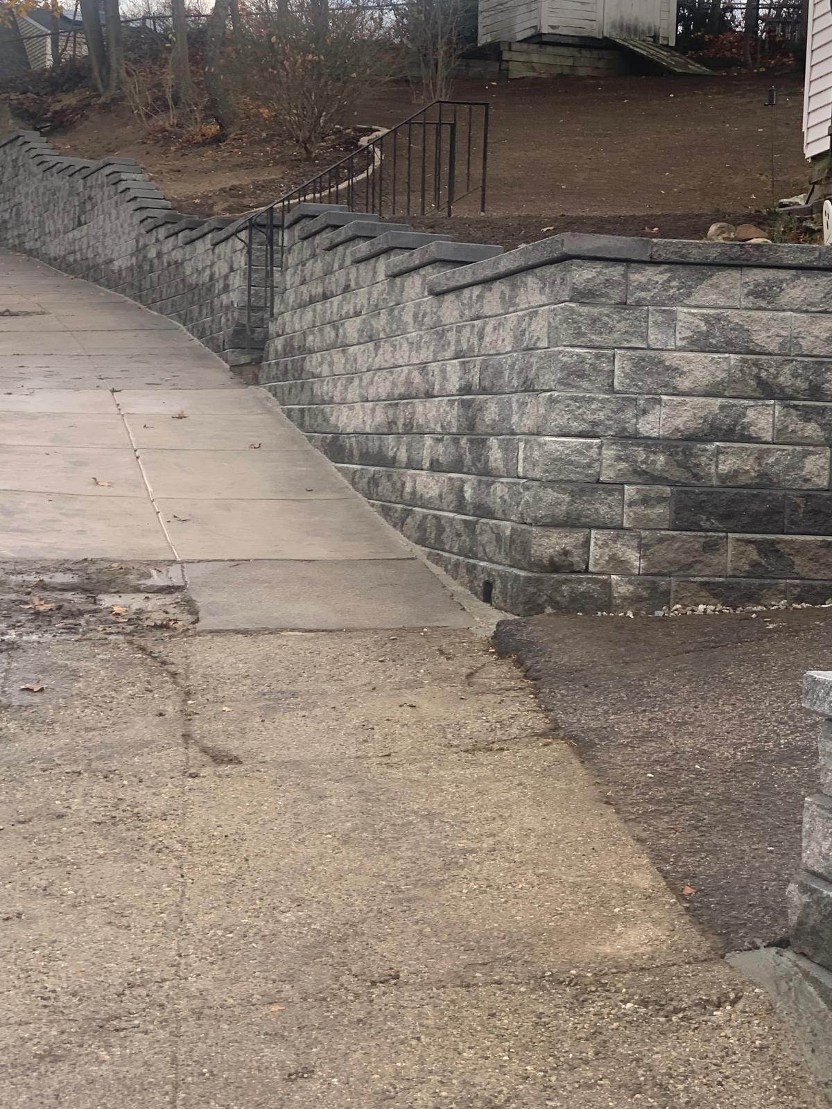 Concrete sidewalk beside a gray stone retaining wall along a sloped walkway outdoors