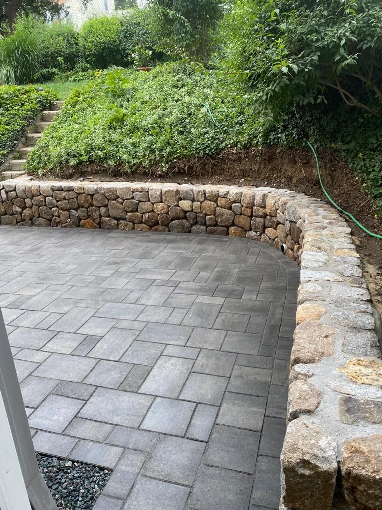 Paved patio beside a curved stone retaining wall and lush green hillside garden.