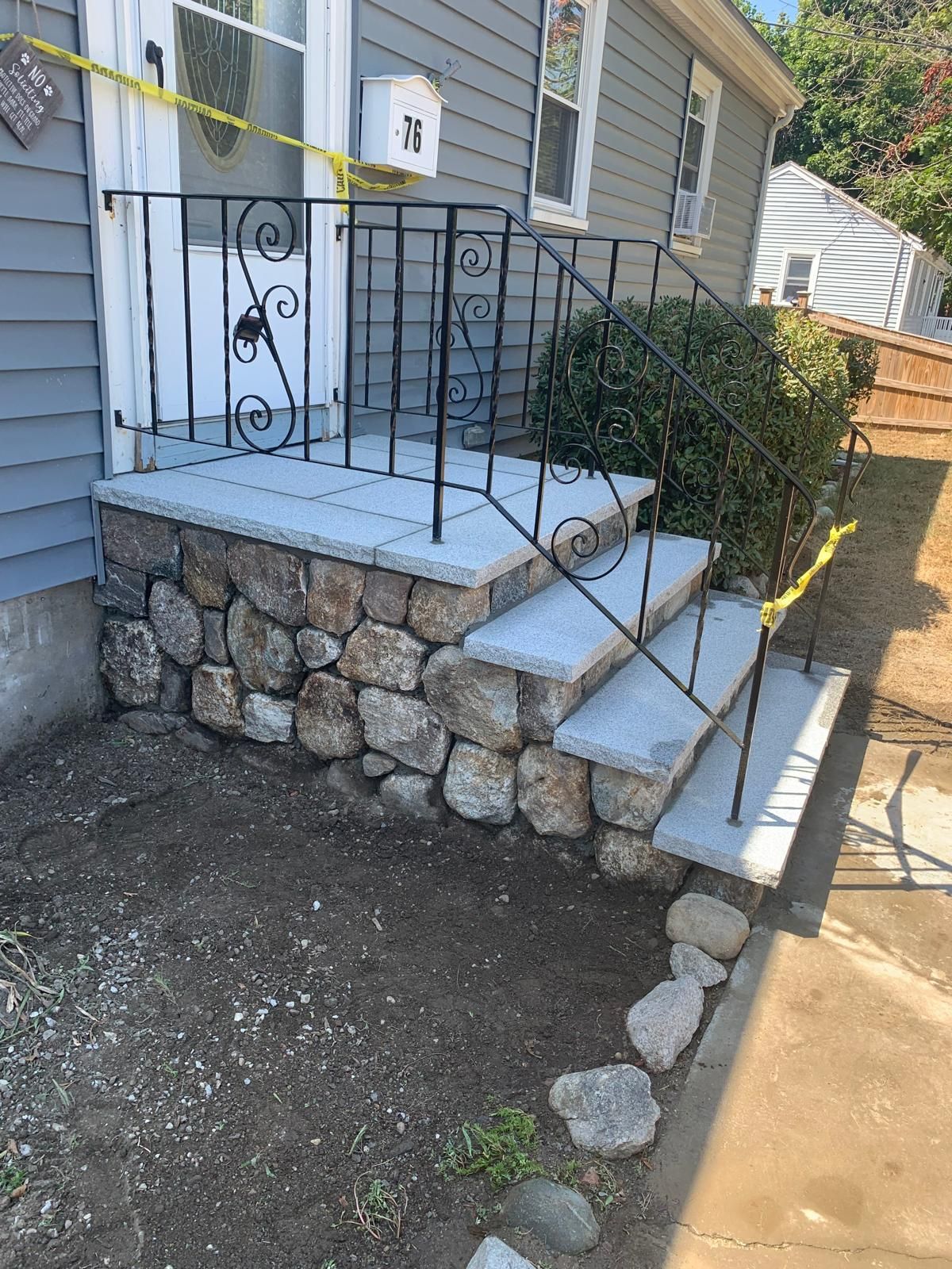 Front steps of a house with stone retaining wall, black railing, and sidewalk beside a sloped gravel yard