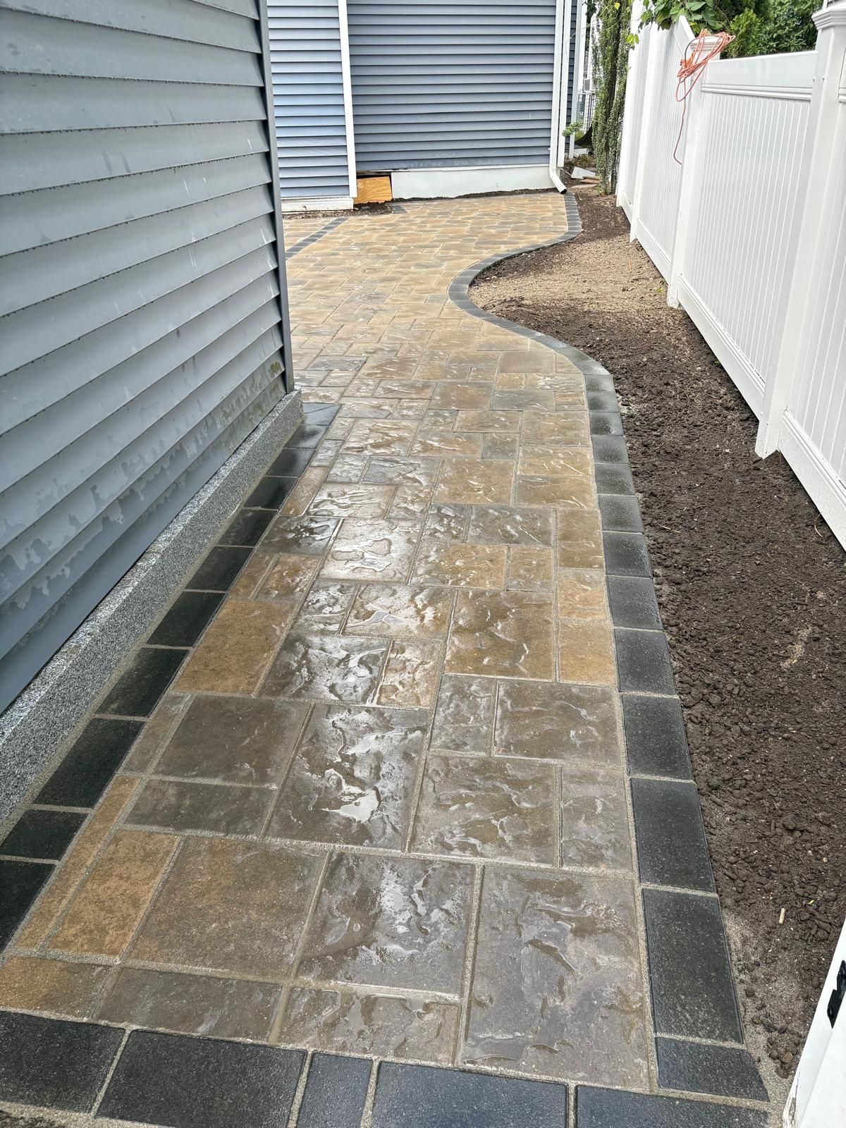 Narrow stone-paved walkway beside a house, with gray siding and a white fence.