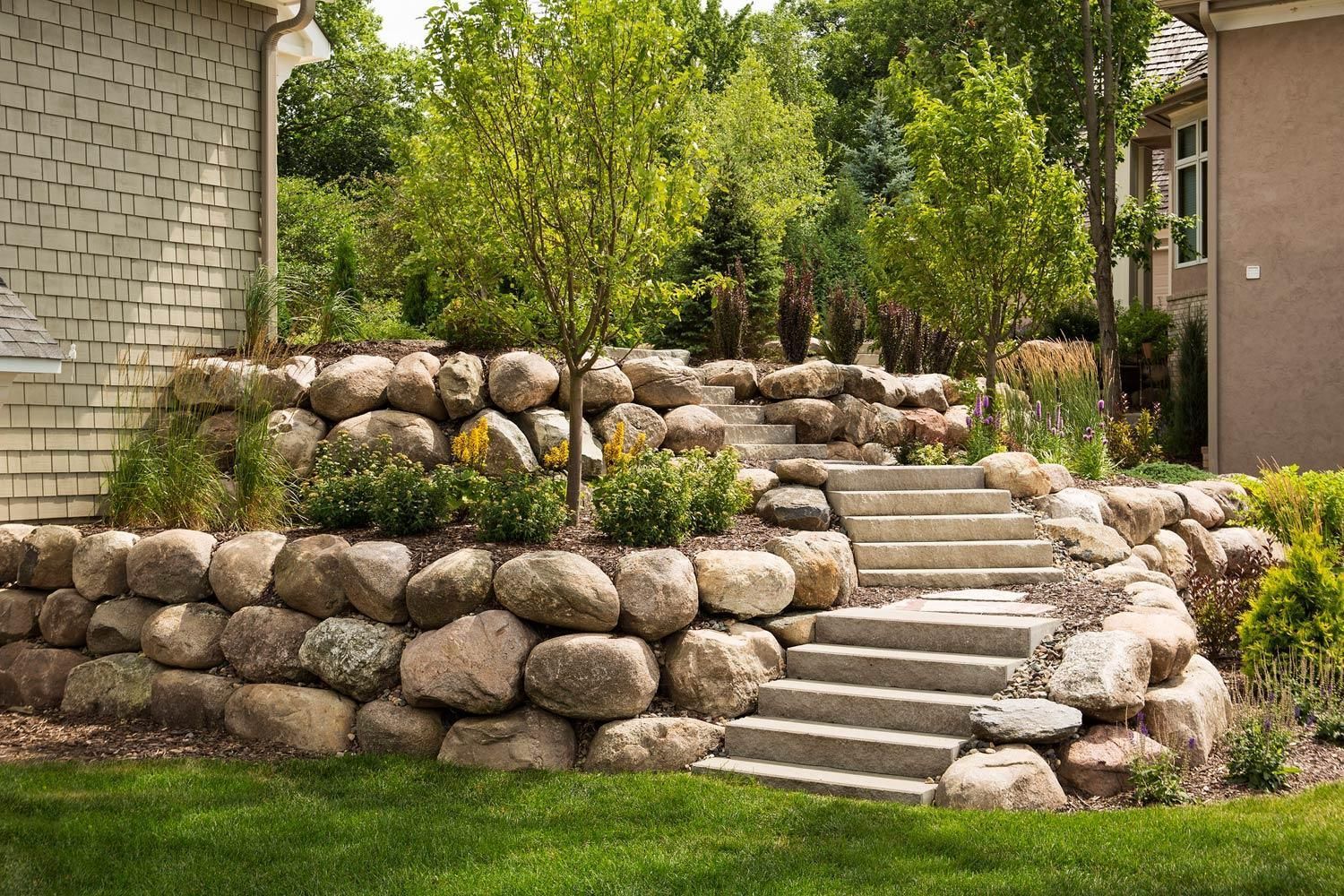 Stone terraced garden steps with stacked rock retaining walls and green shrubs beside buildings