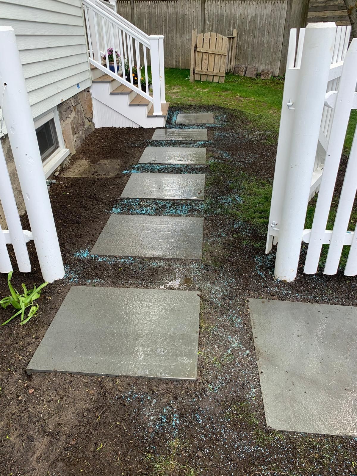 Stepping-stone walkway through a narrow side yard beside a house, bordered by white fencing and damp gravel.