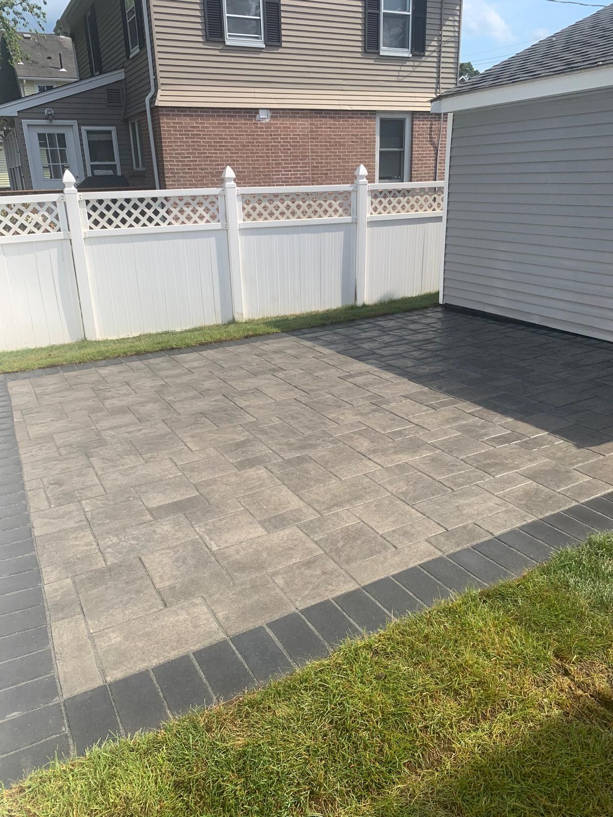 Small paved patio with gray pavers, bordered by dark edging beside a white fence and garage.