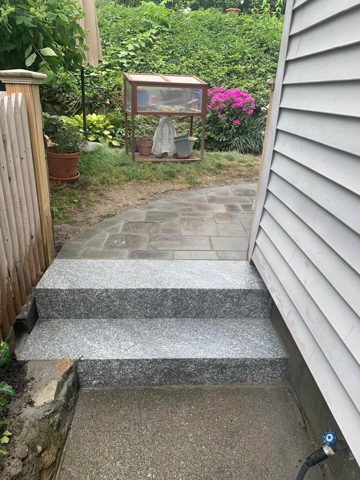Narrow stone steps leading to a small garden path beside a house, with greenery and flowers ahead