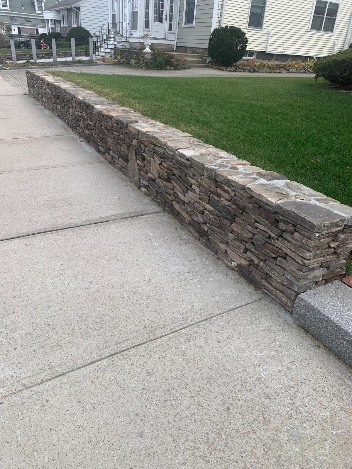 Stone retaining wall along a concrete sidewalk beside a green lawn and house exterior