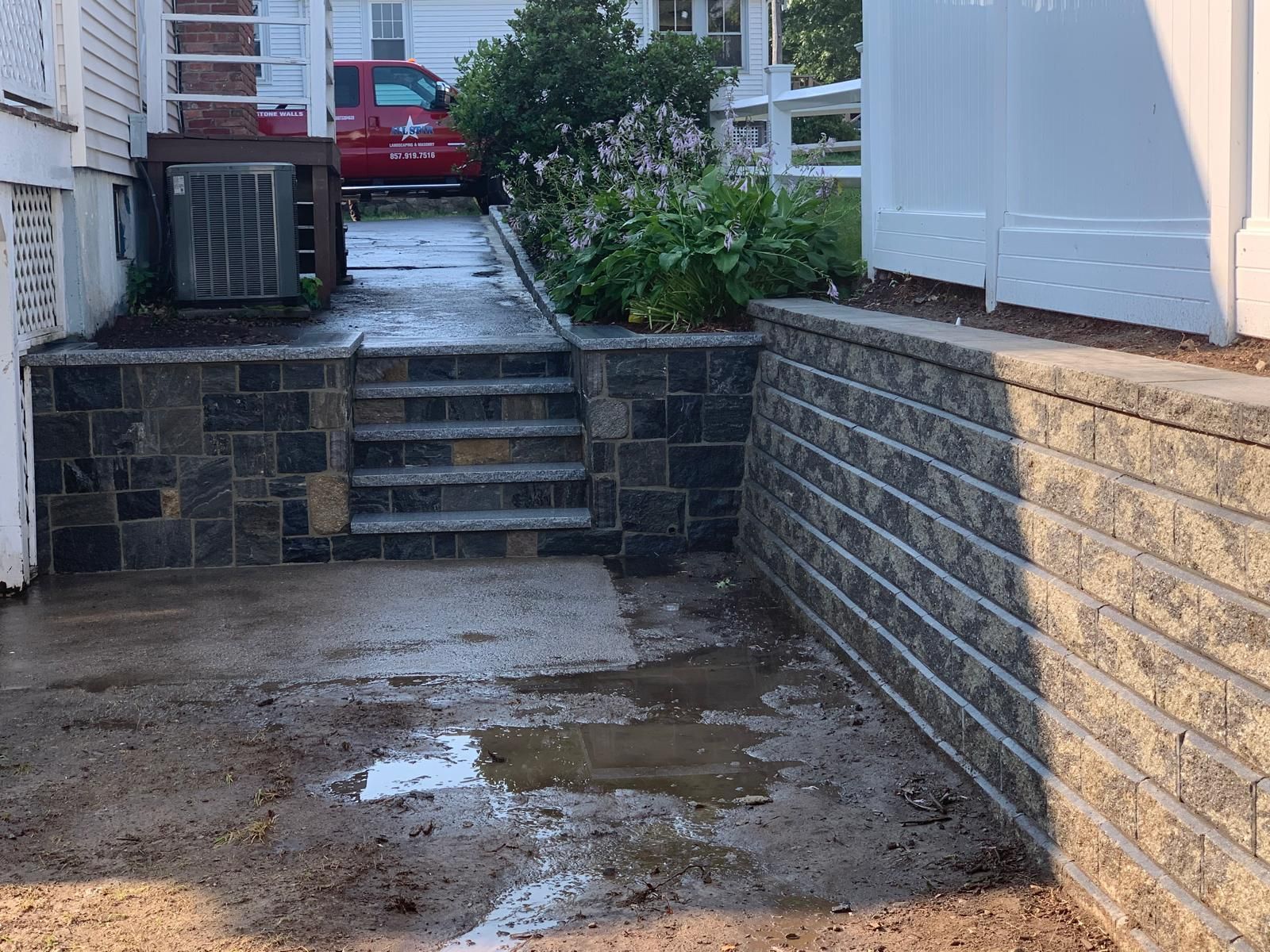 Backyard concrete driveway with steps, stone retaining walls, puddles, and a red car in the distance