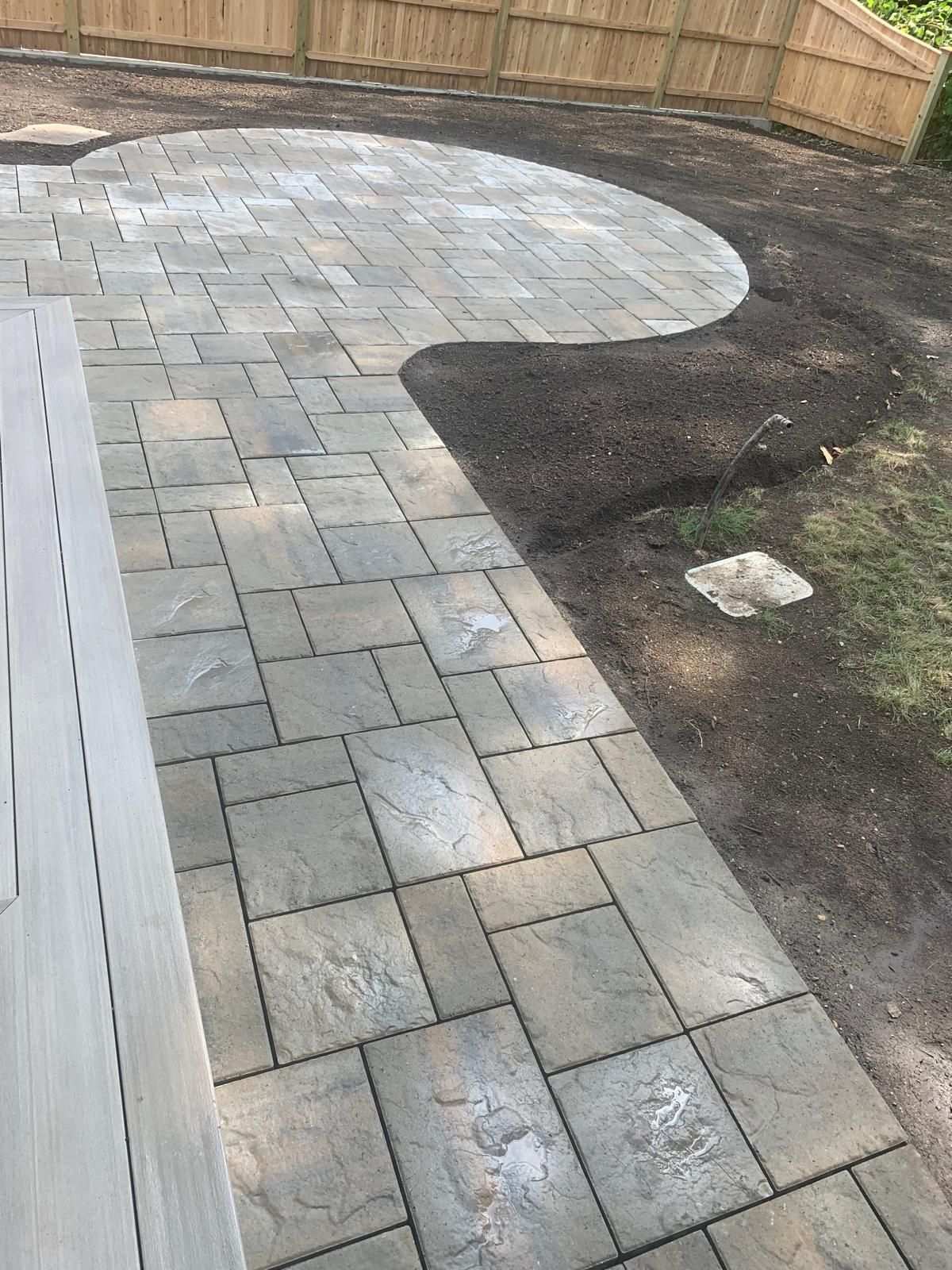 Curved gray paver patio beside a wooden fence and bare soil in a backyard