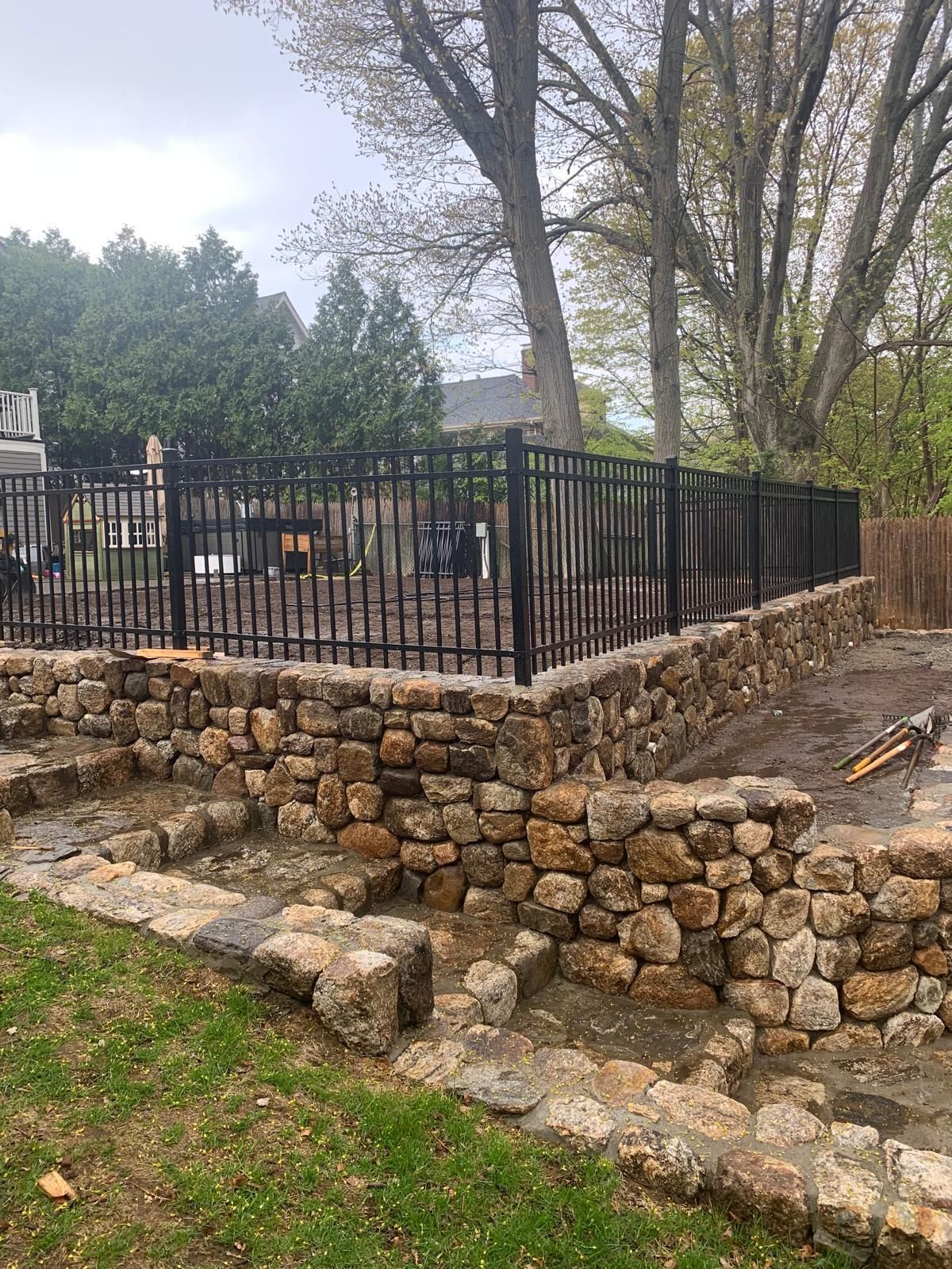 Stone retaining wall with black metal fence on a grassy slope beside trees and a patio area