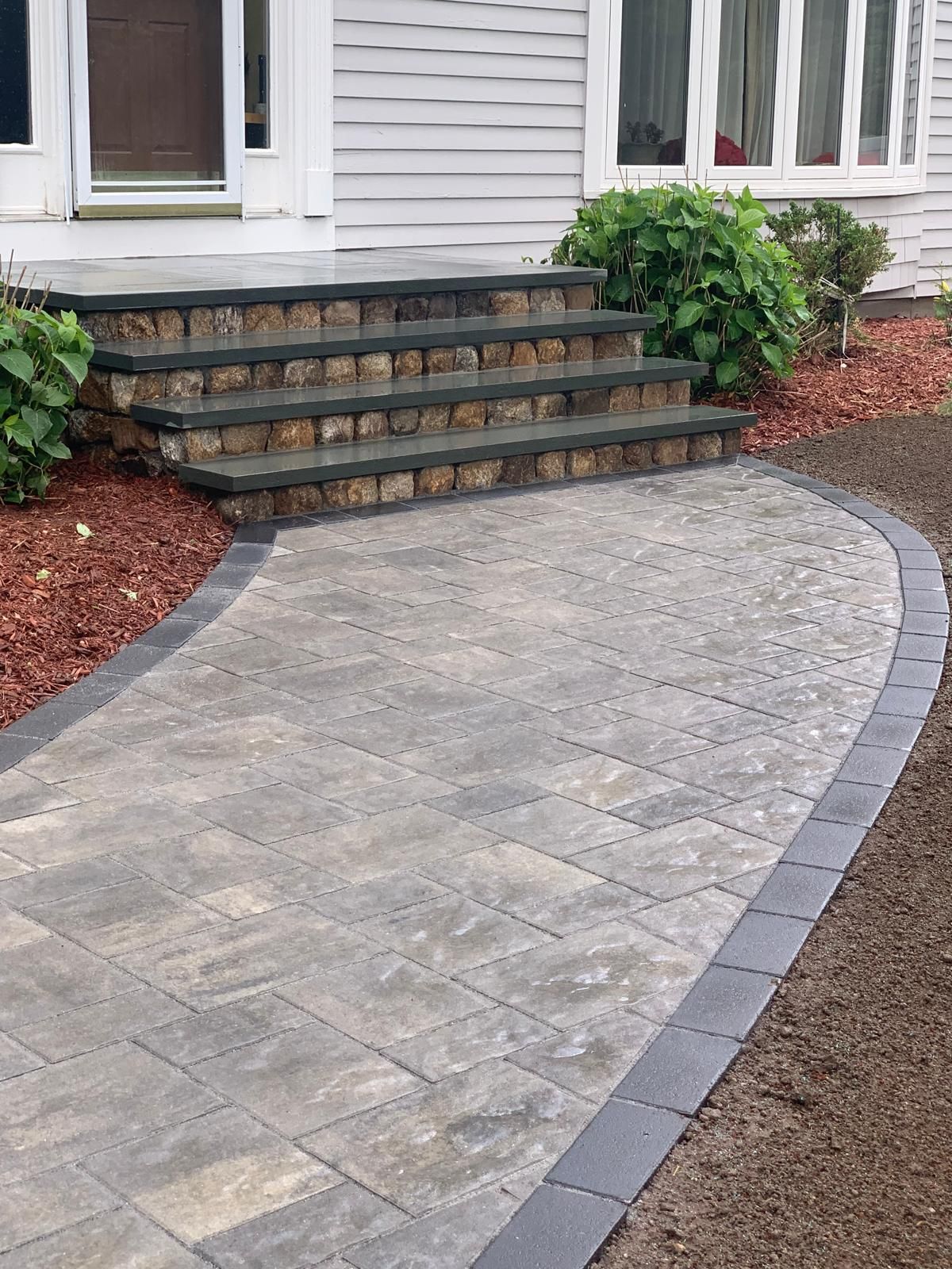 Curved stone-paver walkway leading to front steps and a house entrance with shrubs and mulch