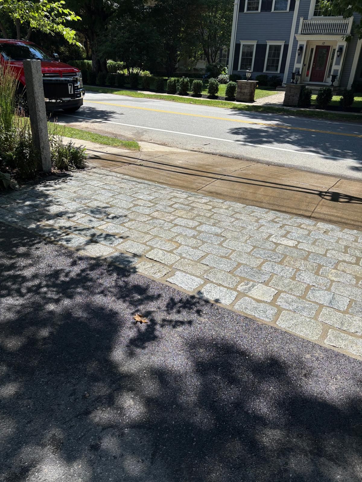 Sunlit driveway and sidewalk in a suburban neighborhood, with a parked red car and house across the street