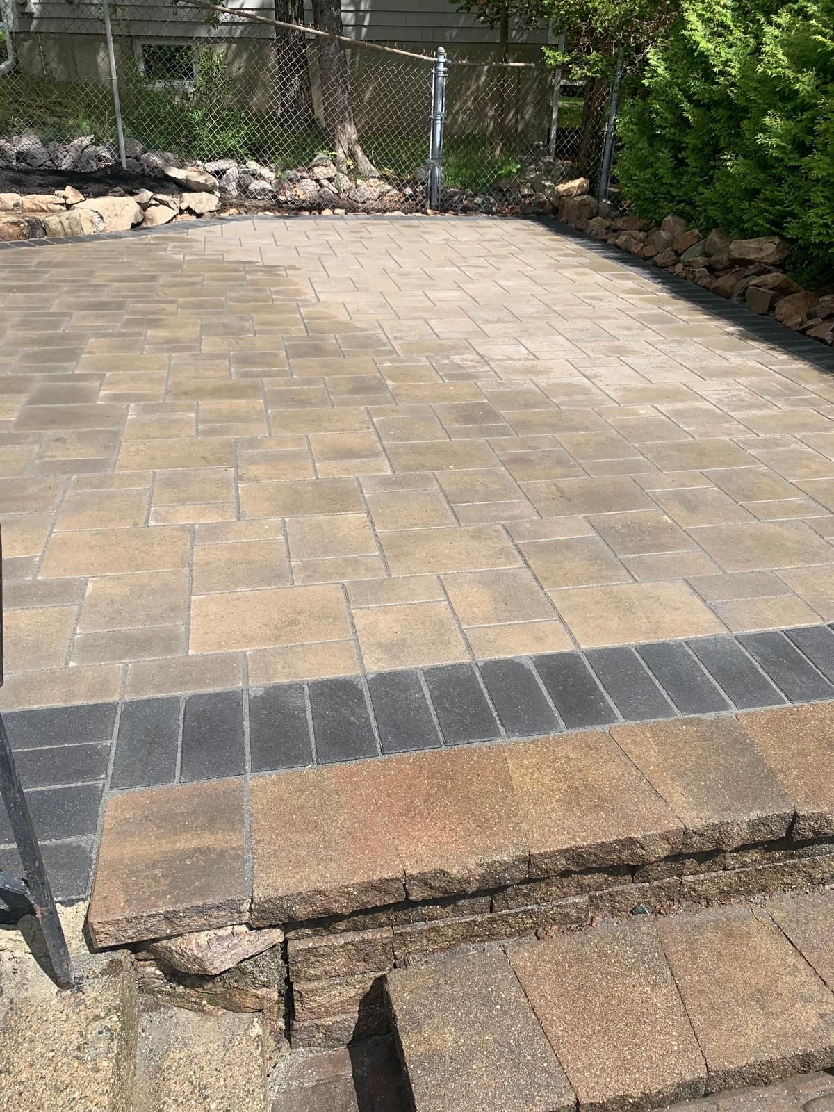 Stone patio with a raised step, bordered by rocks and shrubs in a backyard