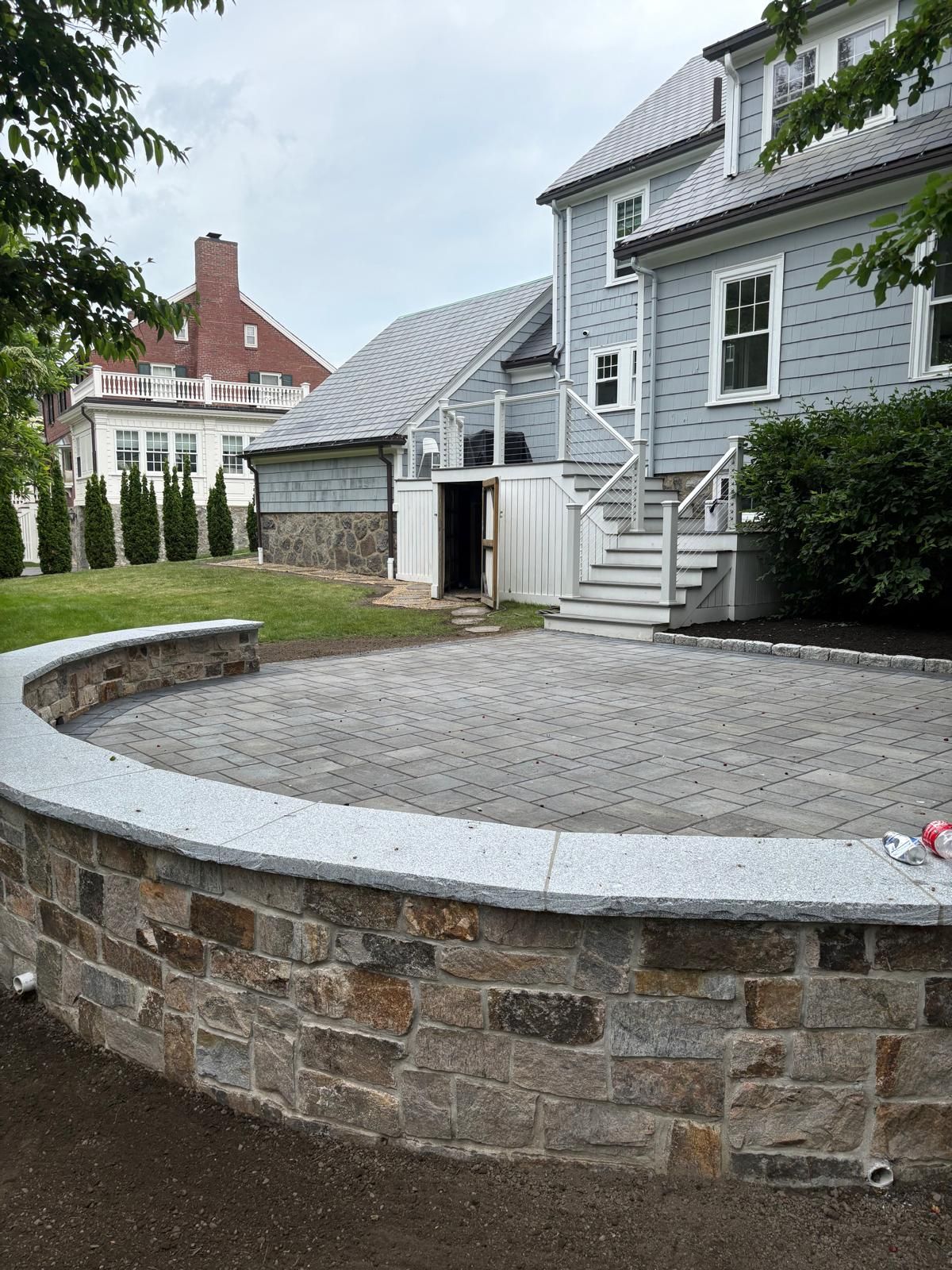 Stone-walled patio beside gray house with steps, gravel ground, and lawn in a residential yard
