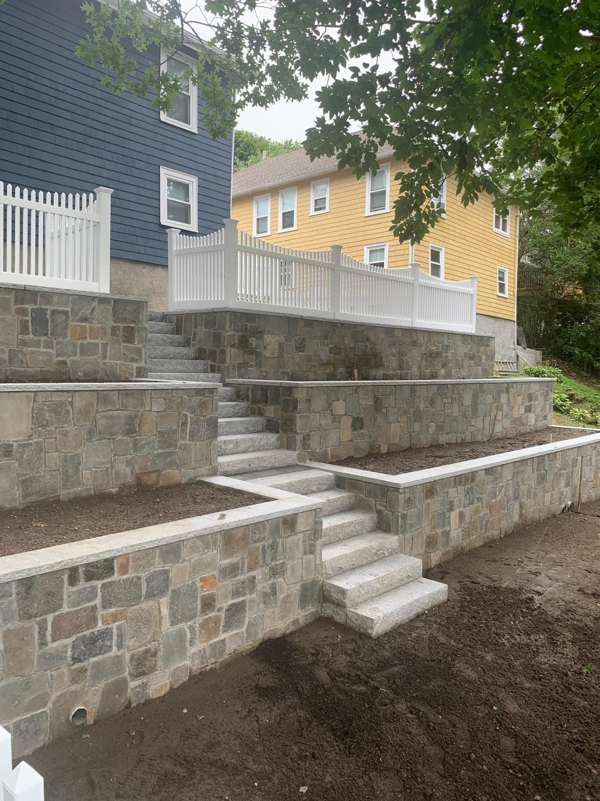 Tiered stone retaining walls with white railings beside blue and yellow houses and a small stairway.