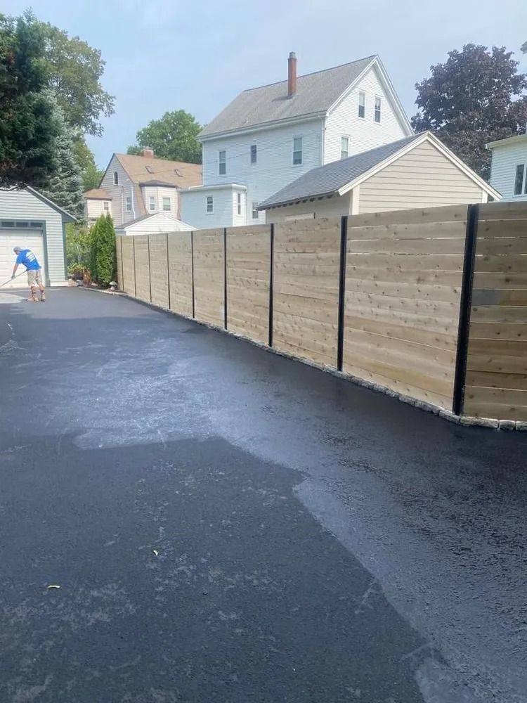 Paved driveway beside a tall wooden fence, with houses and a person near a gate in the background.