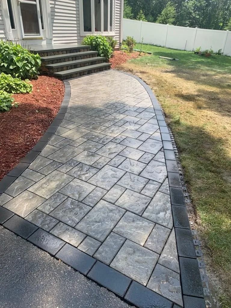 Curved gray paver walkway beside a house, bordered by red mulch and grass, leading to front steps