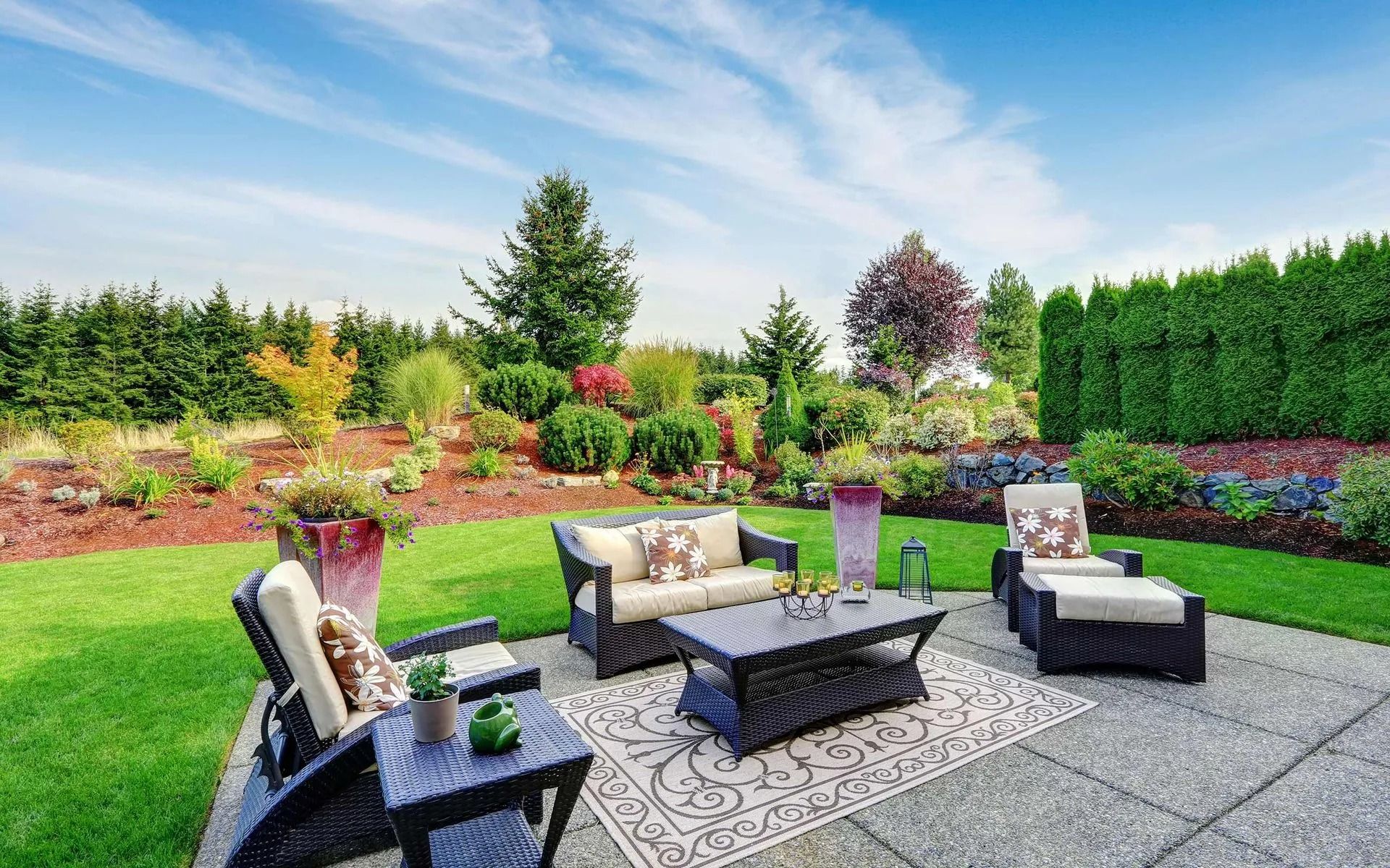 Backyard patio with wicker seating, fire pit, and landscaped garden under a bright sky