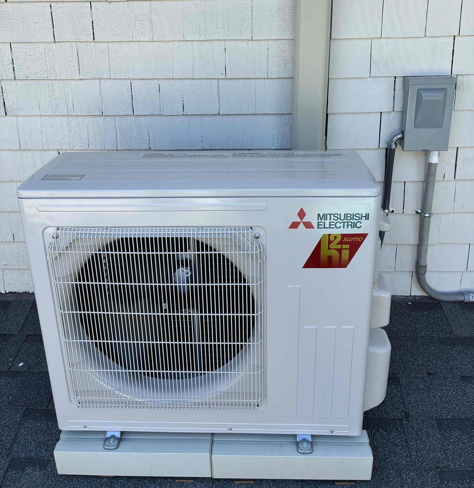 White Mitsubishi Electric mini-split outdoor unit beside a building wall and electrical box
