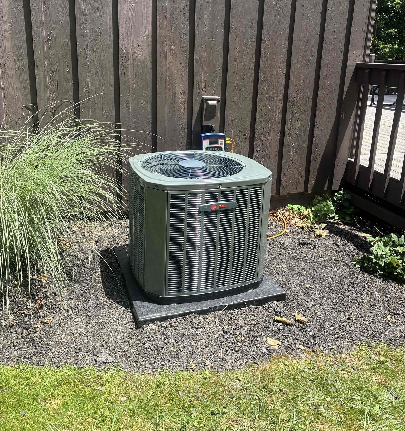 Outdoor air conditioner unit on a gravel pad beside a fence and patio railing, next to ornamental grass.