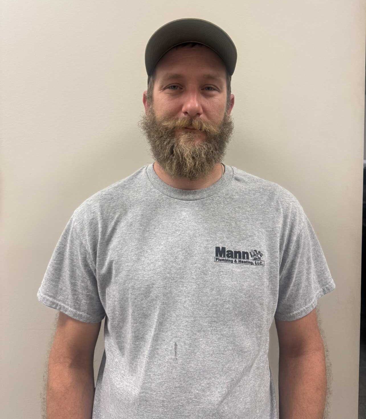 Man with a beard and baseball cap wearing a gray shirt with a logo, standing in front of a neutral wall.